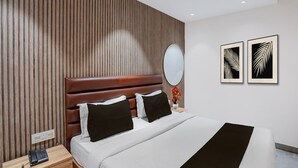 Ein Hotelzimmer mit einer Wand aus Holzlatten, einem braun gepolsterten Kopfteil, einer weißen Bettdecke, zwei schwarzen Kissen, einem Nachttisch mit Telefon und zwei gerahmten Blattformen an der Wand.