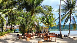 An outdoor seating area with tables and chairs, surrounded by palm trees, overlooking a body of water.