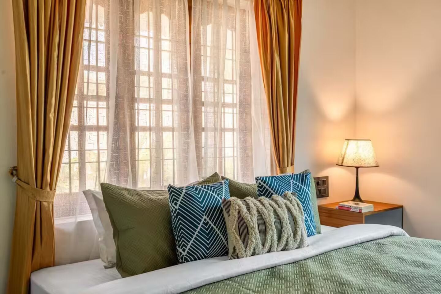 A hotel room with a bed, pillows, a nightstand with a lamp, and sheer curtains.