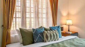 A hotel room with a bed, pillows, a nightstand with a lamp, and sheer curtains.