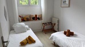 A hotel room with two beds, a window with a view of greenery, a radiator, and a small wooden chair. - Le MARMARE Vacation Rental (Caussou)