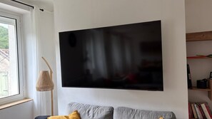 A room with a large black artwork on the wall, a window with white curtains, a woven lamp, a grey sofa with cushions, and a bookshelf with books and a black object. - Le MARMARE Vacation Rental (Caussou)