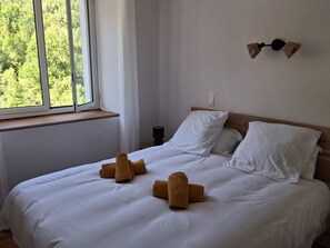 A hotel room with a bed, white linens, a window with a view of greenery, and a wall-mounted light fixture. - Le MARMARE Vacation Rental (Caussou)