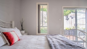 Ein Schlafzimmer mit einem Bett, einem Nachttisch mit Pflanze, einem Fenster und einer Tür zu einem Außenbereich.