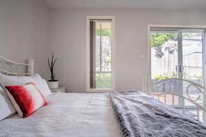 Un dormitorio con una cama, una mesita de noche con una planta, una ventana y una puerta que da a un espacio exterior.