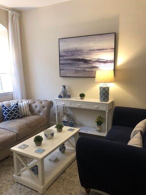 A living room with a beige sofa, a dark armchair, a white coffee table, a white side table with decorative items, a wall-mounted lamp, and a painting of a landscape. - Pet-Friendly Woodland Escape, 30 acres, Turriff. (Turriff)