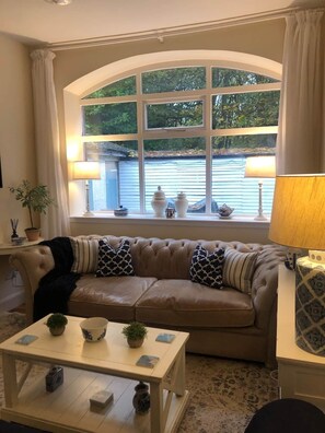 A cozy living room with a large arched window, a leather sofa with patterned cushions, a small table with decorative items, and a lamp on a side table. - Pet-Friendly Woodland Escape, 30 acres, Turriff. (Turriff)
