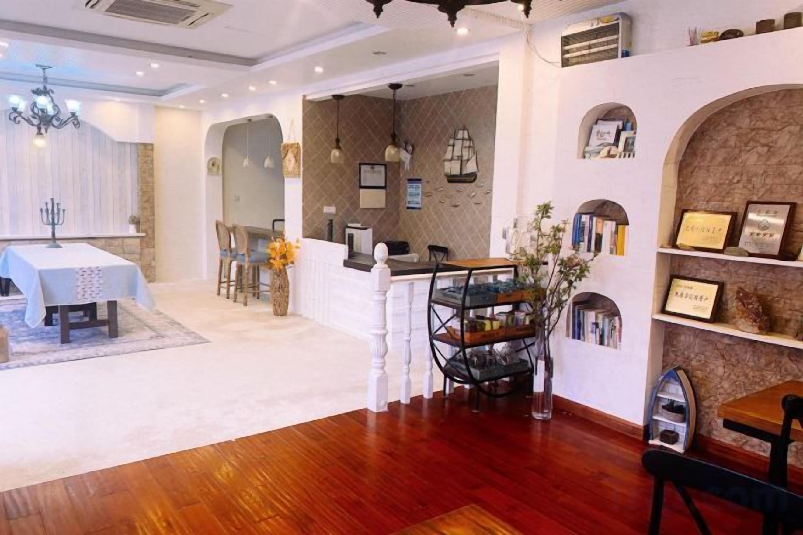 A hotel lobby with a reception desk, a dining area with a table and chairs, and a wall with shelves displaying framed items.
