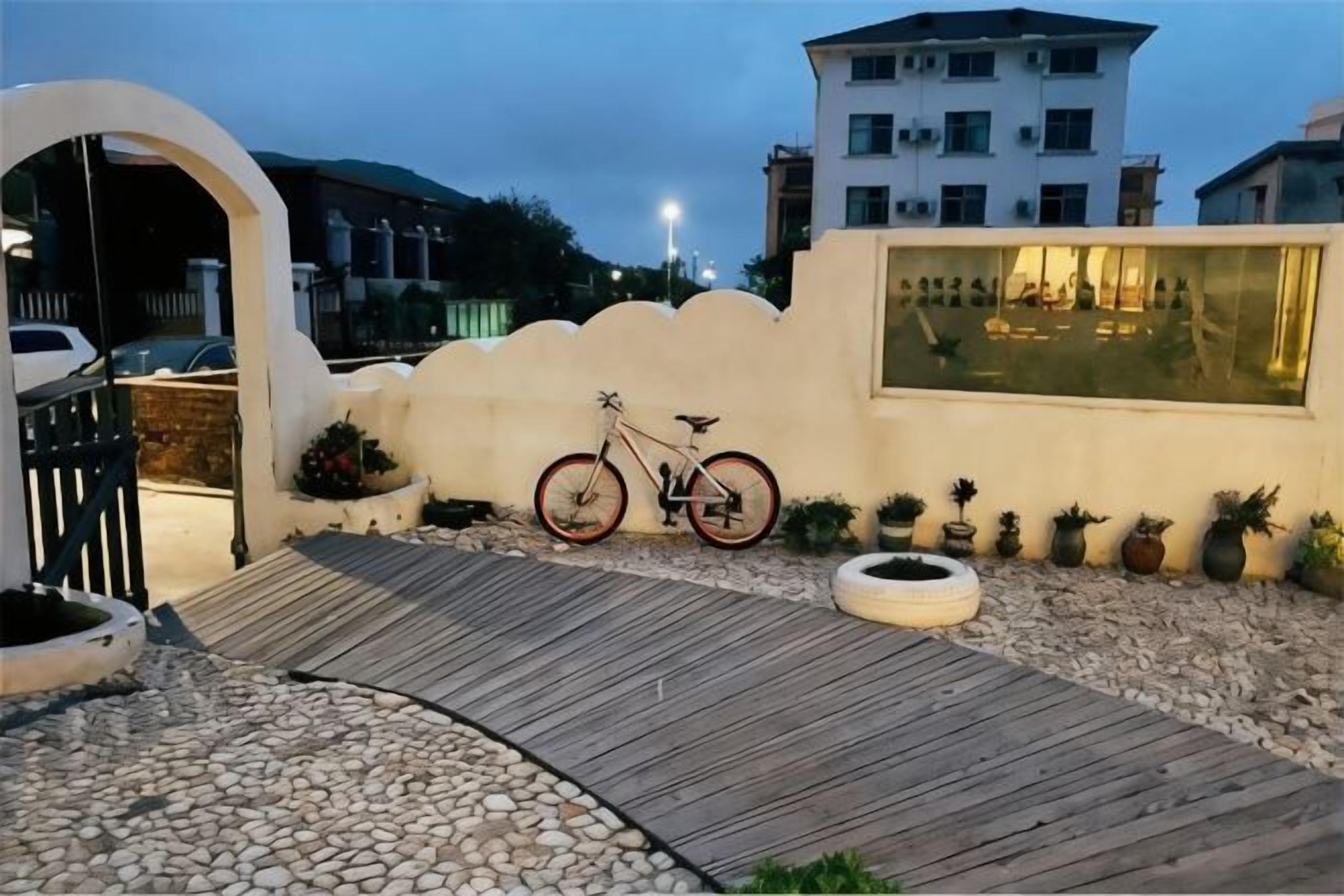 A paved entrance with a bicycle, a white wall with a window, and a building in the background.