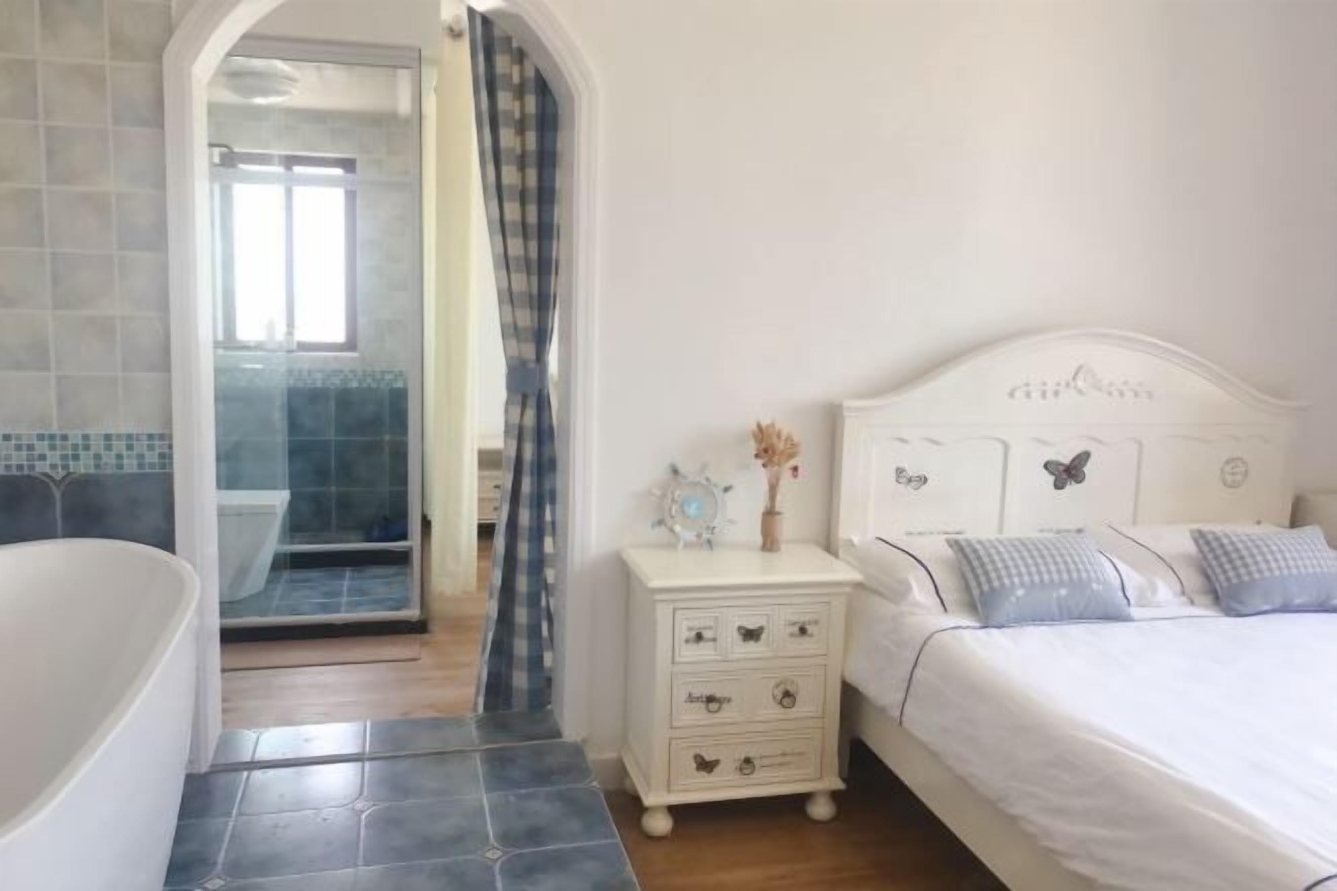 A bedroom with a white bed, a nightstand, a dresser, and a bathroom with a bathtub and shower.