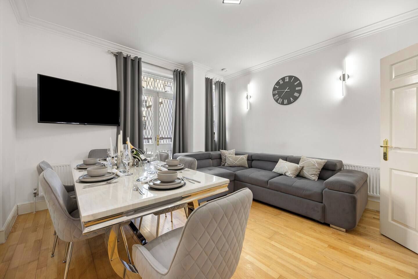 Una sala de estar moderna con zona de comedor, un sofá gris, un televisor de pantalla plana y un reloj de pared.