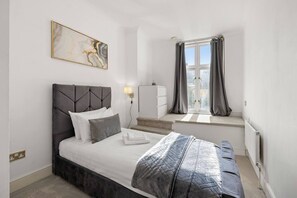 A bedroom with a bed, a nightstand, a window with curtains, a radiator, and a wall-mounted light. - Luxury Marylebone Flat | 2 Bed 2 Bath (Greater London)