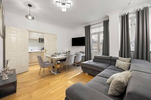 Ett modernt vardagsrum med en grå soffa, en matplats med stolar, en platt-TV som är monterad på väggen och ett kök som syns genom en öppen dörr.