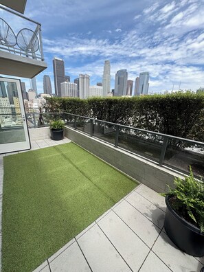 A rooftop terrace with artificial grass, potted plants, and a city skyline view.