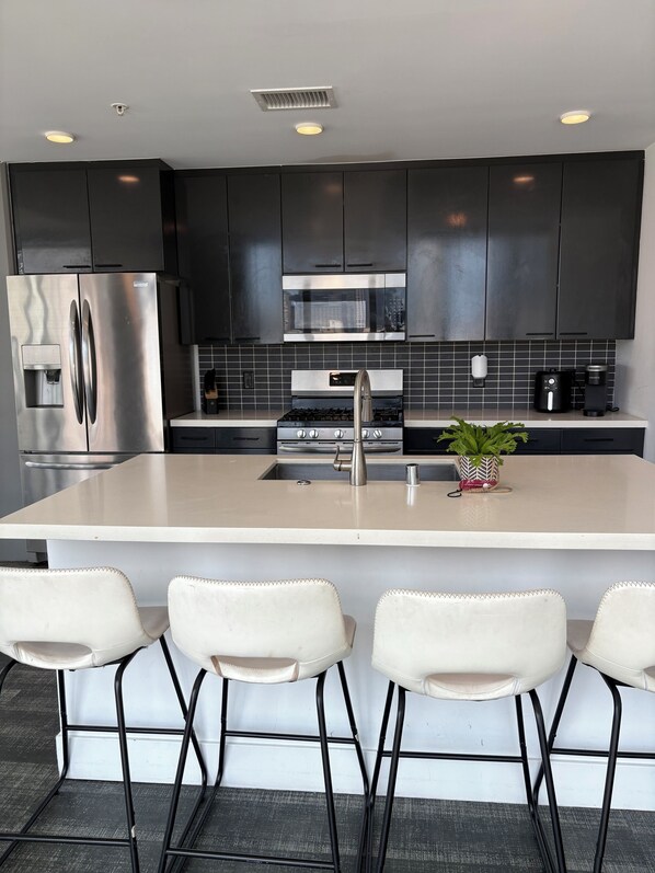 A modern kitchen with a stainless steel refrigerator, a built-in microwave, a gas stove, and a sink. There are four white bar stools in front of the kitchen island.