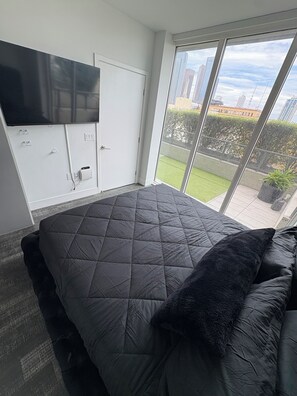 A bedroom with a large bed, a television mounted on the wall, and a sliding glass door leading to a balcony with a city view.