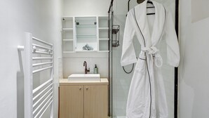 A modern bathroom with a glass shower enclosure, a wooden cabinet, a white sink, and a robe hanging on a hook. - Stylish 1BR in the Heart of Le Marais (Paris)