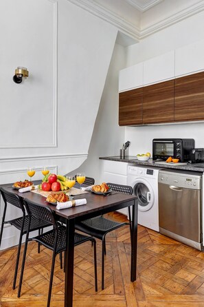 A modern kitchen with white cabinetry, a built-in oven, microwave, and dishwasher. There is a dining table with chairs and a breakfast setting. - Stylish 1BR in the Heart of Le Marais (Paris)