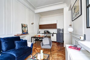 A modern living room with a blue sofa, a dining table with chairs, a kitchen area with a refrigerator and microwave, and a hallway with a framed picture. - Stylish 1BR in the Heart of Le Marais (Paris)