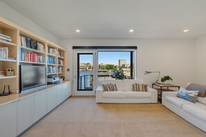 Un salon moderne avec un canapé blanc, un téléviseur à écran plat, une bibliothèque et une vue sur le paysage urbain, visible à travers une grande fenêtre.