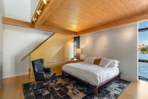 Ein Schlafzimmer mit einem Bett, einem Sessel, einem Nachttisch, einer Lampe und Blick auf das Wasser.