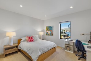 Une chambre moderne avec un lit, des lampes de chevet, un bureau et une fenêtre donnant sur un immeuble et l’eau.