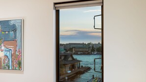 A room with a window overlooking a waterfront, a framed painting on the wall, and a stack of books on a table. - Stunning Waterfront! Tom Kundig-Designed, Dock, Fire Pit & Bay Views (Seattle)