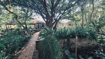 A garden pathway leading to a gazebo with a thatched roof, surrounded by lush greenery and a brick wall.