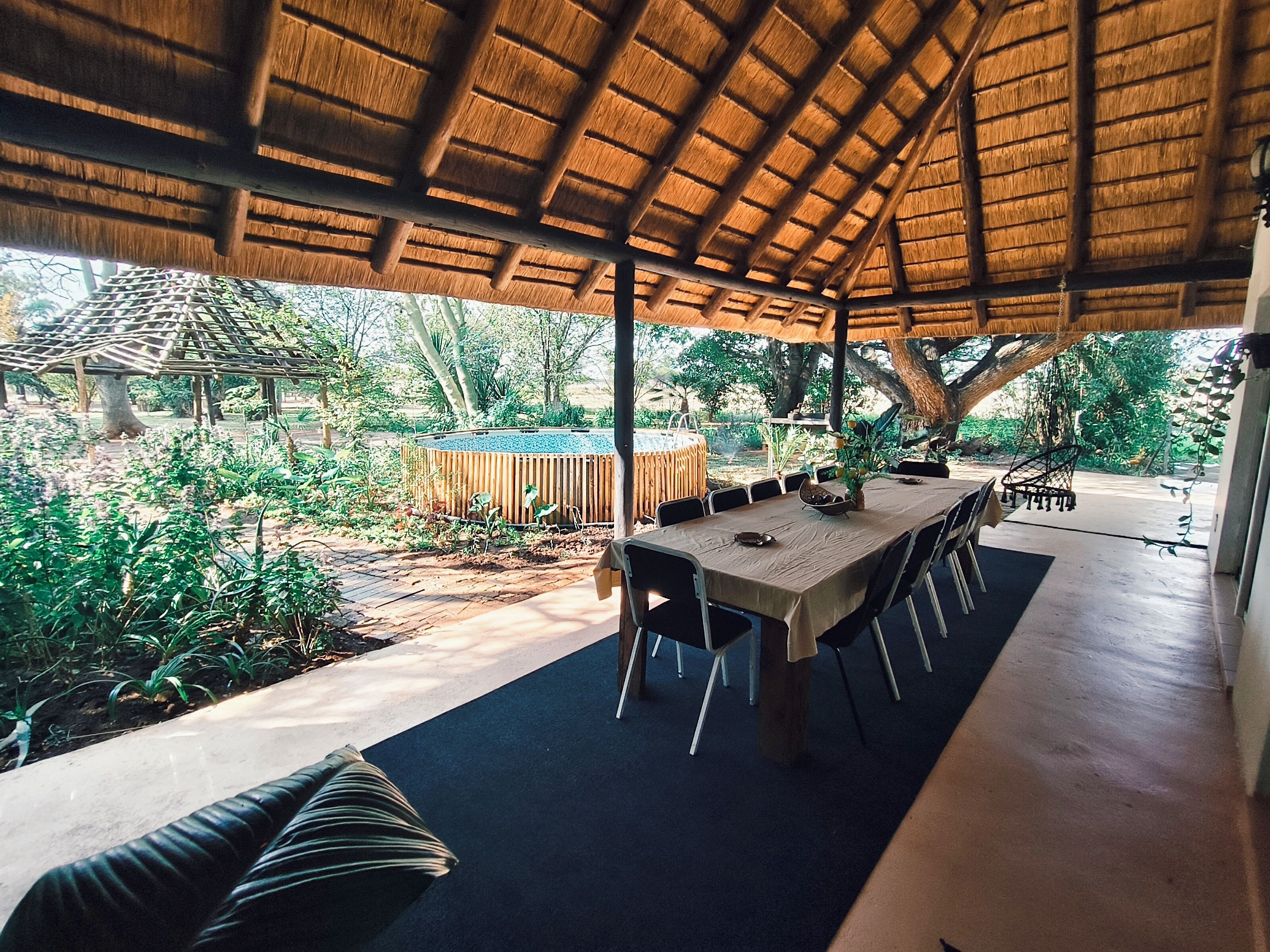 An outdoor dining area with a thatched roof, surrounded by greenery and a pool.