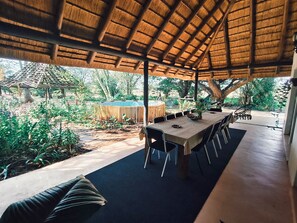 Un espacio de comedor al aire libre con techo de paja, rodeado de vegetación y una piscina.