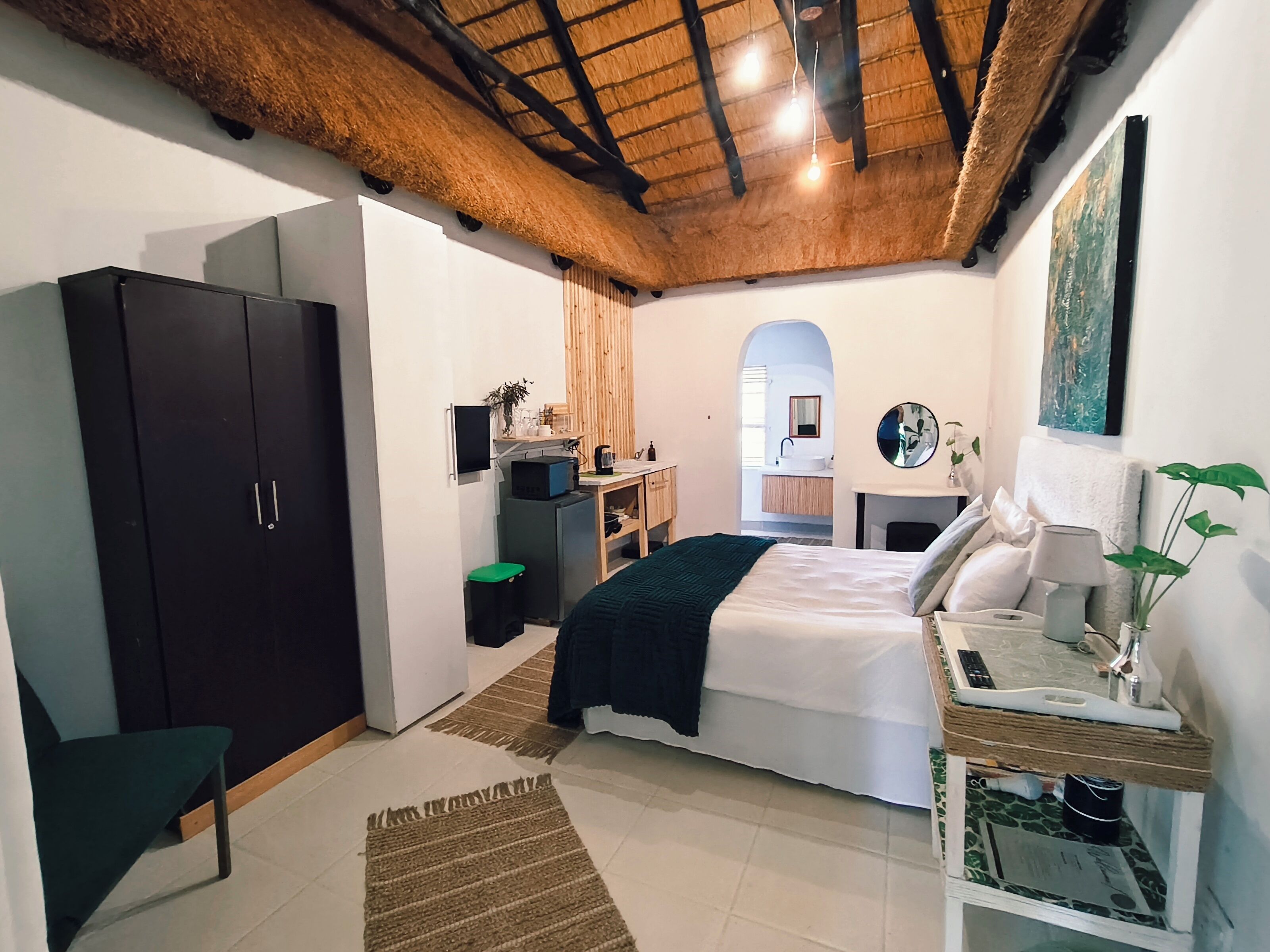 Un dormitorio con cama, mesita de noche y armario. Hay un escritorio con silla y un espejo. La habitación tiene techo de madera y una ventana.