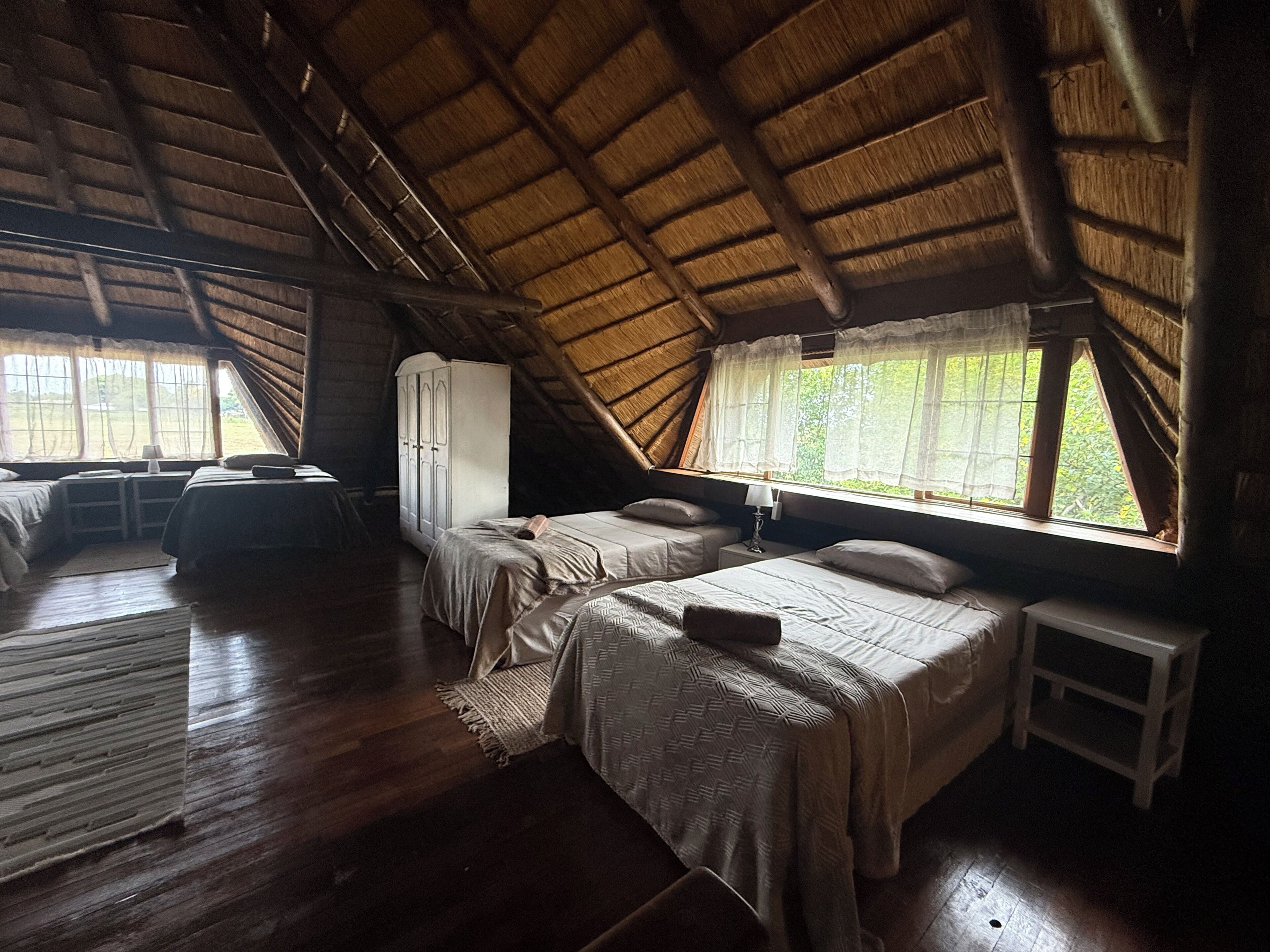 A room with a sloped ceiling, wooden floors, two beds, a wardrobe, and a window with curtains.