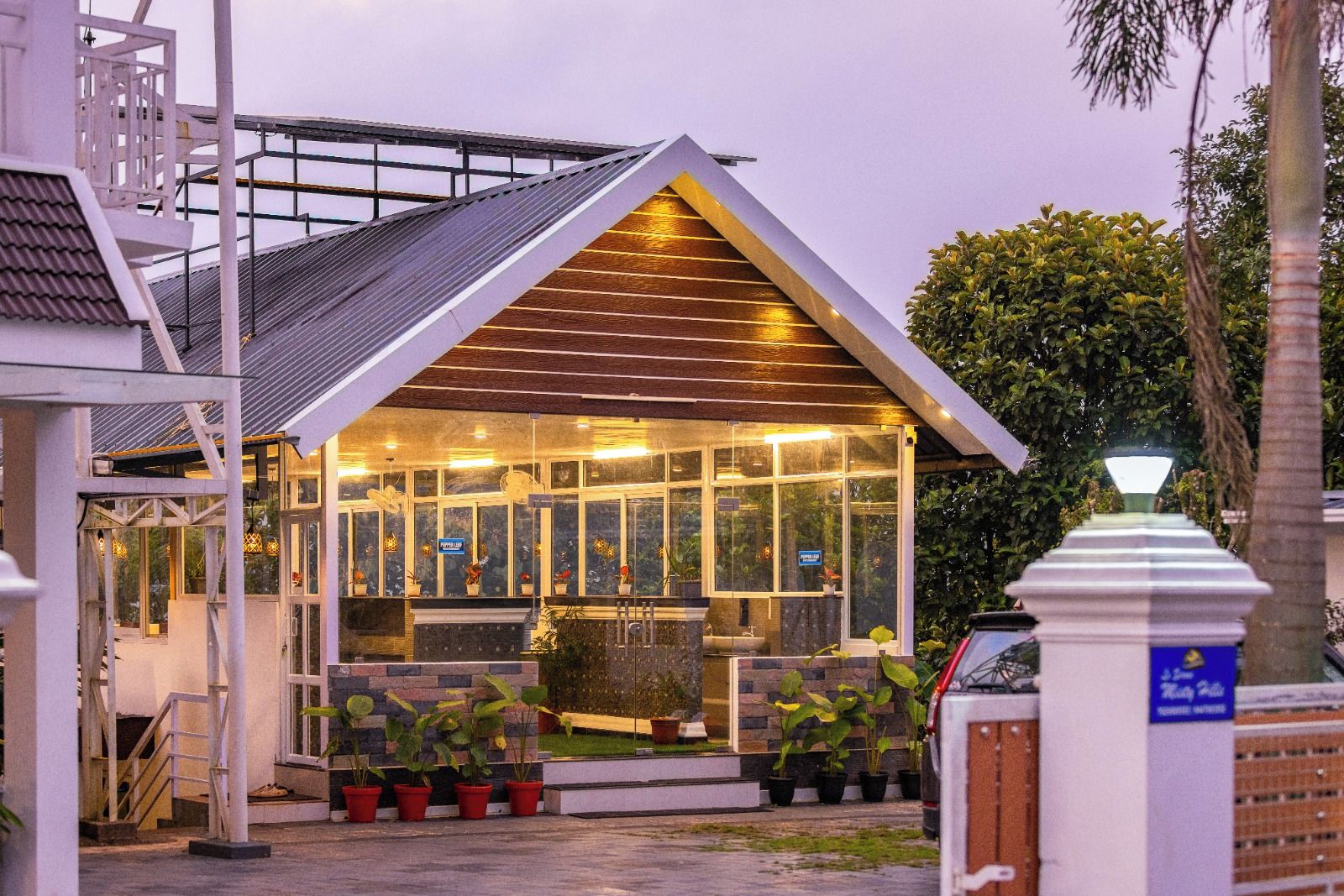 Um edifício moderno com fachada de vidro, telhado inclinado de madeira e área de entrada pavimentada com plantas em vasos.