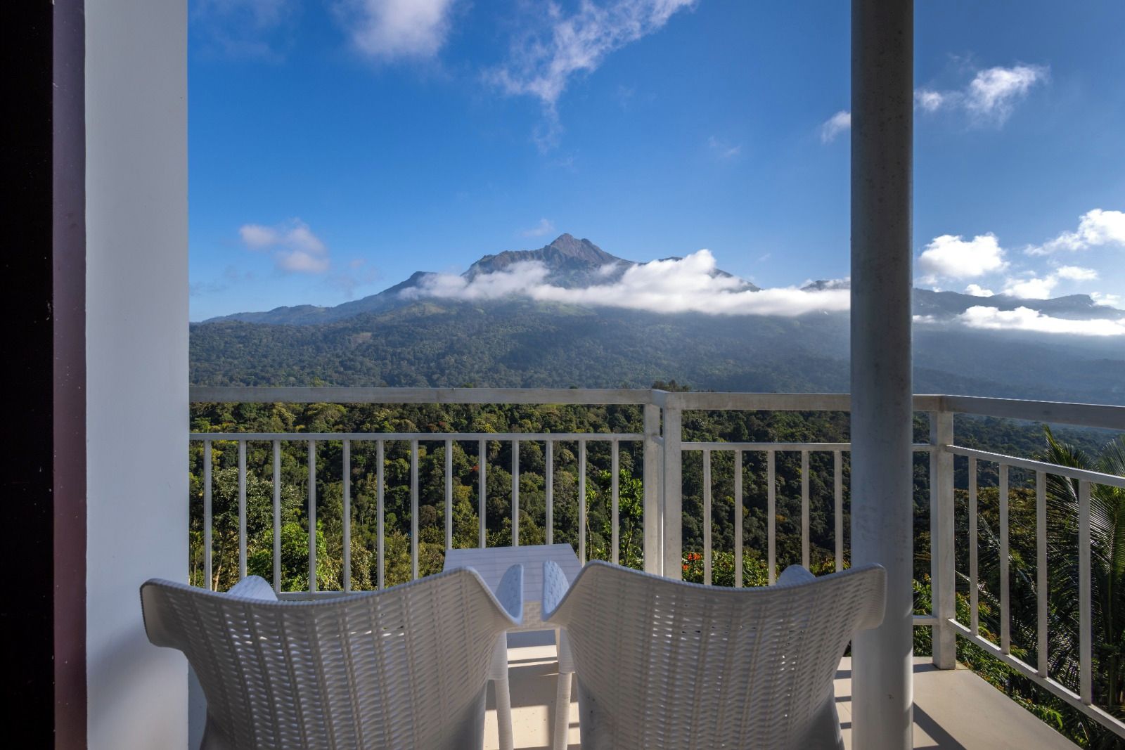 A balcony with wicker chairs, overlooking a mountainous landscape with a prominent peak and lush greenery.