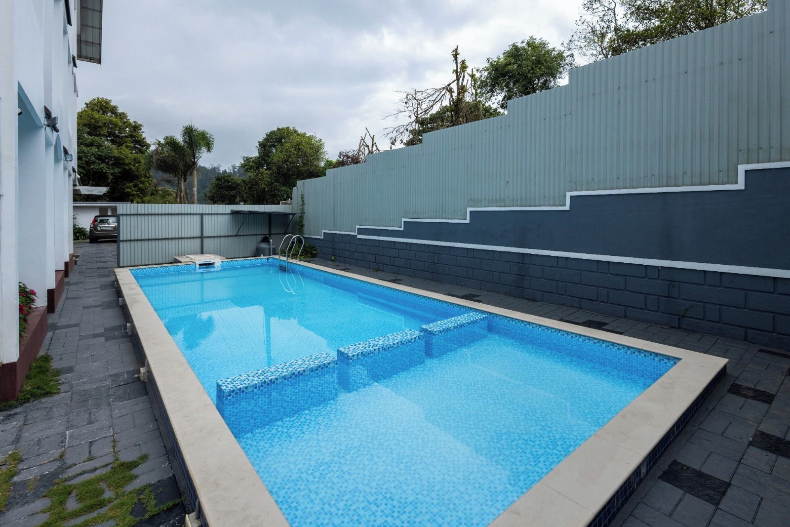 Una piscina rectangular con borde de azulejos, rodeada por un sendero empedrado y un muro bajo.
