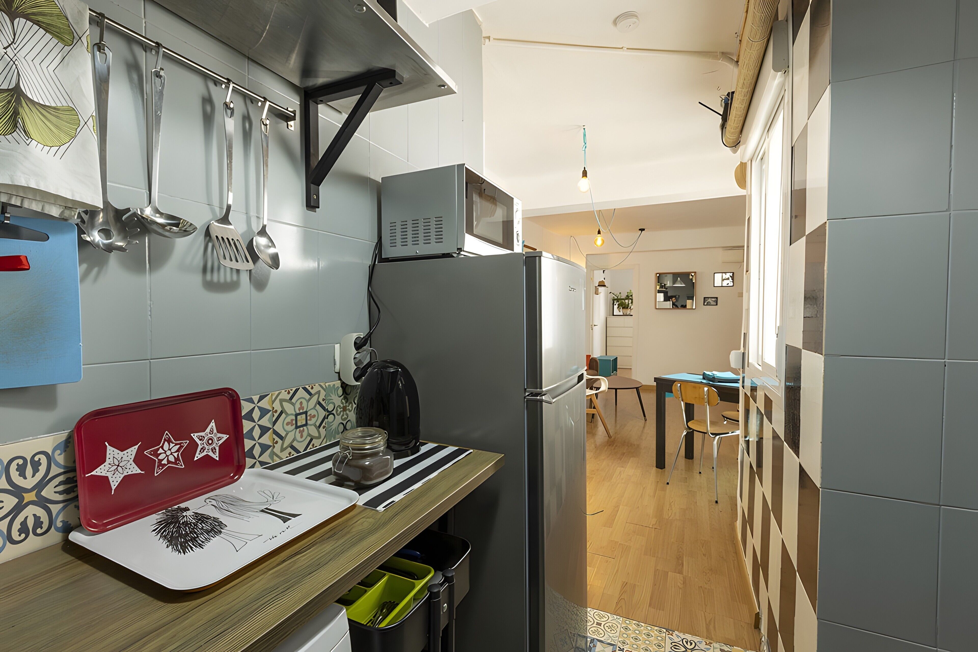 Eine kleine Küche mit Kühlschrank, Wasserkocher und Mikrowelle. Es gibt eine Holz Arbeitsplatte mit einem roten Tablett und einem schwarzen Wasserkocher. Die Küche hat eine geflieste Spritzschutzwand und ein Fenster.