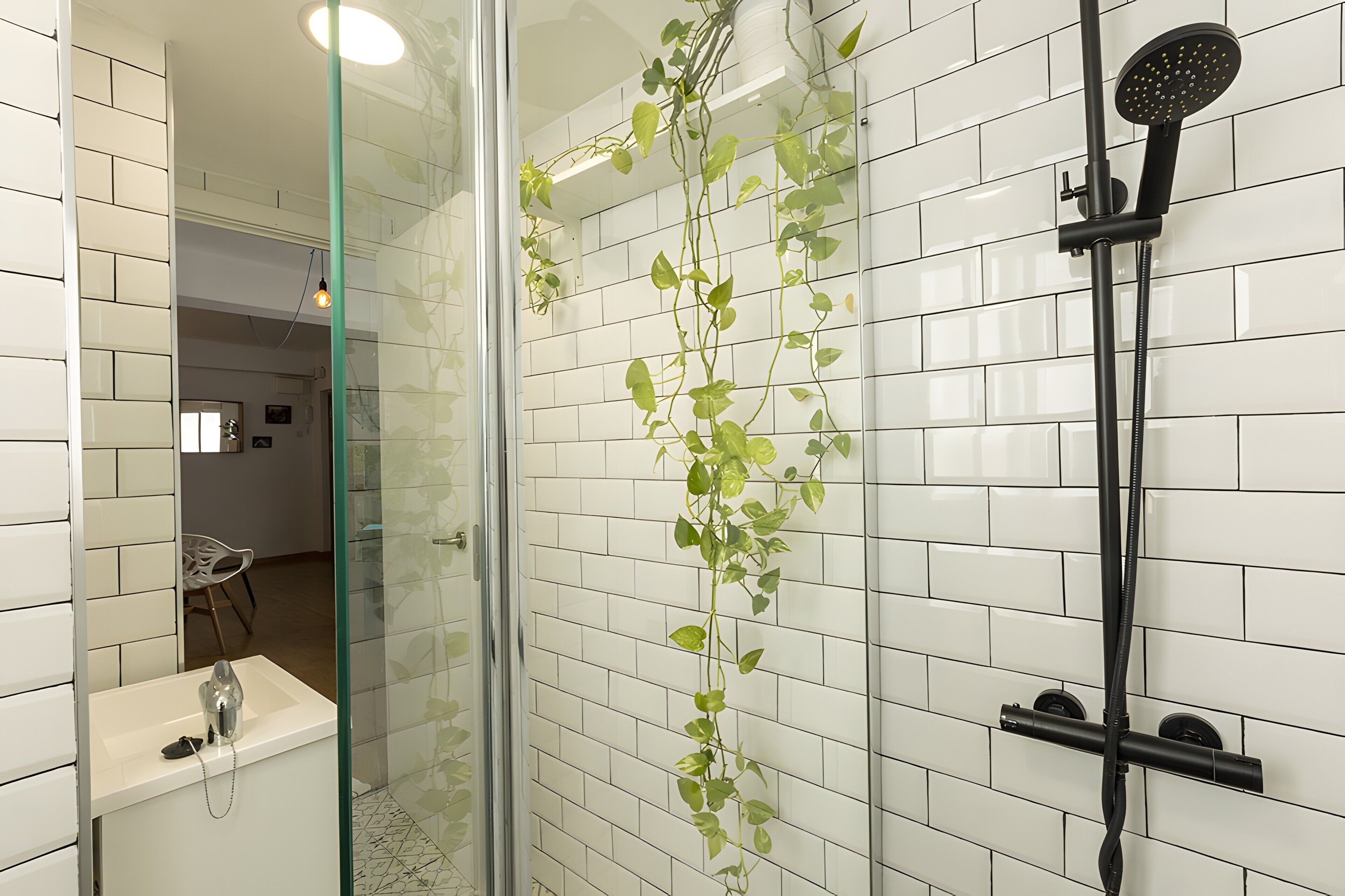 Un baño moderno con paredes de azulejos blancos, cabina de ducha de vidrio, ducha de mano y una planta en maceta.