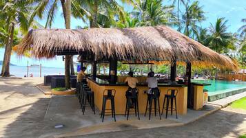 A beachside bar with a thatched roof, outdoor seating, and a view of the ocean.