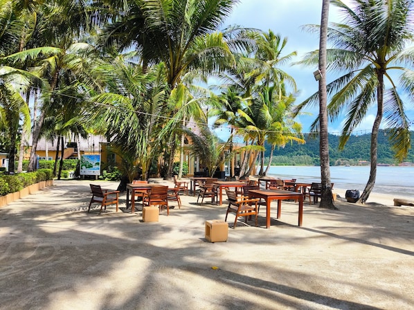 An outdoor seating area with tables and chairs, surrounded by palm trees, overlooking a body of water.