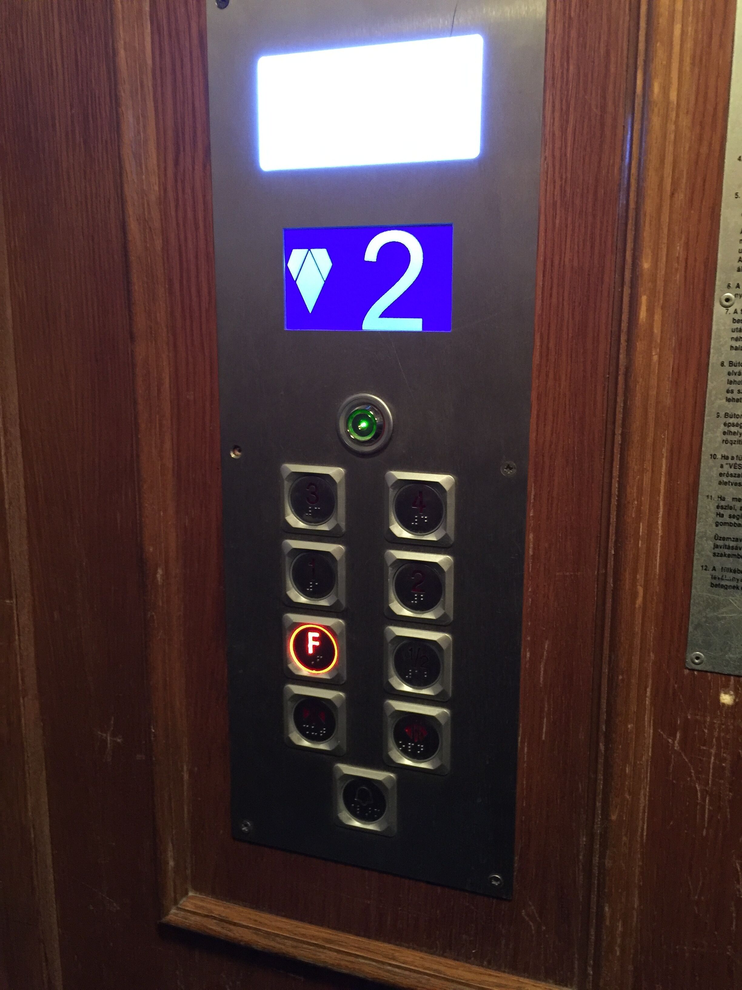 En kontrollpanel för hissen med en digital display som visar våning 2, en grön knapp och en röd knapp för nödstopp.