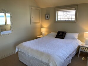 A neatly made bed with a white comforter and pillows, a bedside table with a lamp, a framed picture on the wall, and a window with blinds. - Luxury beachfront villa perfectly located in Ahuriri Napier (Napier)