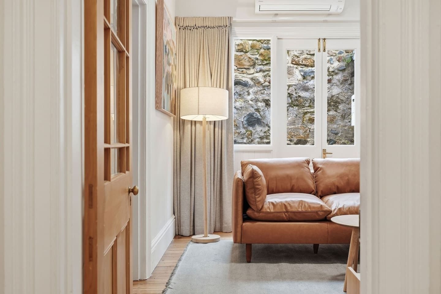 A cozy room with a leather sofa, a lamp, and a view of a stone wall through a window.