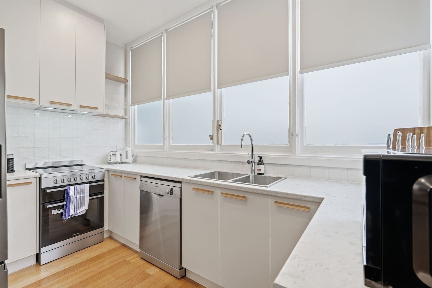 A modern kitchen with white cabinetry, a built-in oven, a dishwasher, and a large window with blinds.