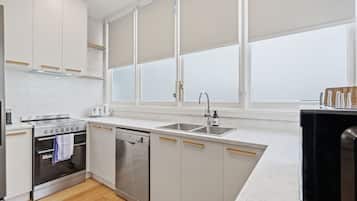 A modern kitchen with white cabinetry, a built-in oven, a dishwasher, and a large window with blinds.