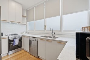 Eine moderne Küche mit weißen Schränken, einem Backofen, einer Spülmaschine und einem großen Fenster mit Jalousien.