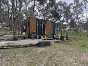 A modern, compact mobile home with a wooden exterior and a black door, surrounded by a grassy area with trees.