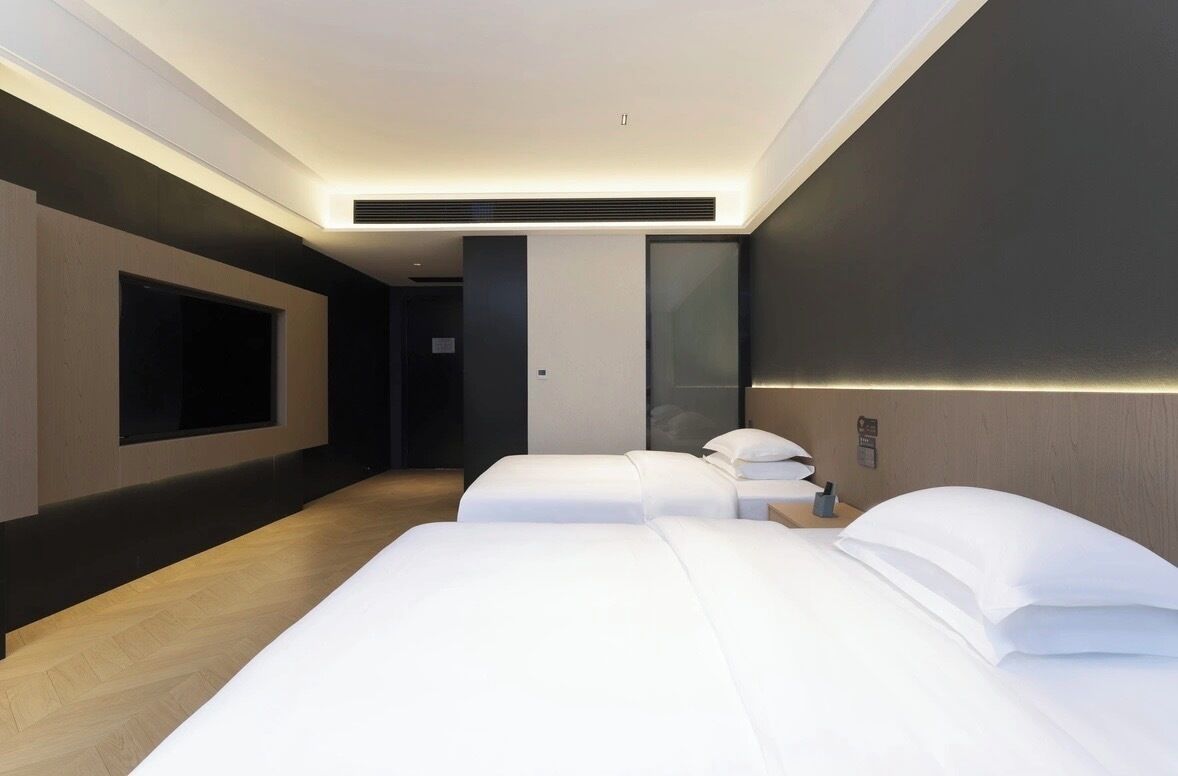 A modern hotel room with two beds, a large flat-screen TV, and a minimalist design.
