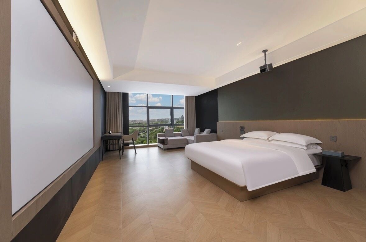 Una habitación de hotel moderna con una cama grande, una zona de estar con sofá y sillón, y un amplio ventanal con vista a la ciudad.