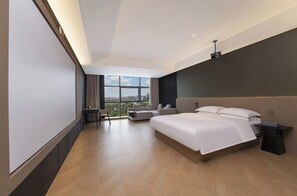 A modern hotel room with a large bed, a seating area with a sofa and armchair, and a large window offering a city view.