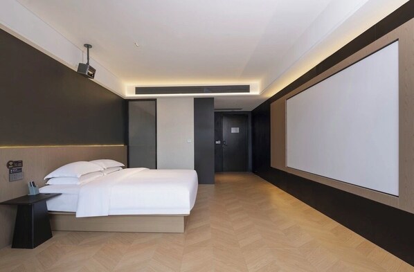 A modern hotel room with a bed, a bedside table, a projector screen, and a large wall-mounted screen.